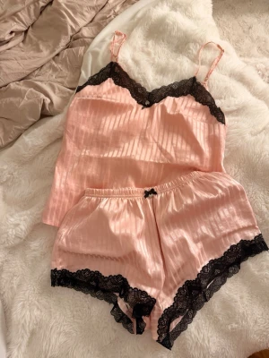 Rosa satinpyjamas med spetsdetaljer - Söt pyjamas i ljusrosa satin med tunna axelband och svarta spetsdetaljer vid halsringning och benslut. Setet består av ett linne och shorts med elastisk midja och liten rosett framtill. Perfekt för mysiga kvällar hemma.
