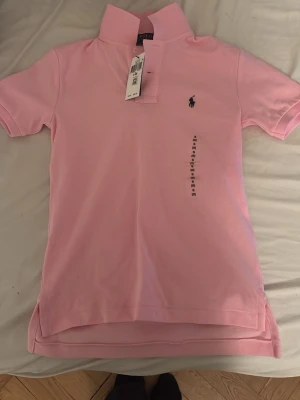 Rosa pikétröja från Polo Ralph Lauren - Klassisk rosa pikétröja från Polo Ralph Lauren med korta ärmar och den ikoniska broderade loggan på bröstet. Tröjan har krage, knappslå och är tillverkad i mjuk bomull. Perfekt för en avslappnad men ändå stilren look.