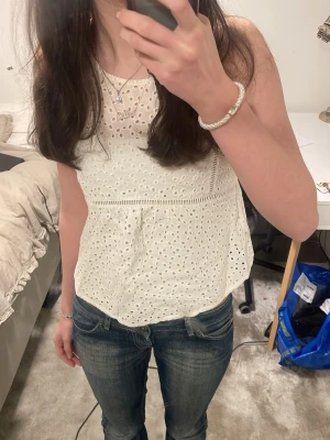 Vit broderad linnetopp i bomull - Söt vit ärmlös linnetopp med broderade detaljer och hålmönster. Toppen har en luftig passform och är tillverkad i bomull, perfekt för varma dagar. Styla med jeans eller kjol för en fräsch look.