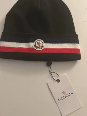 Svart Moncler mössa - Snygg svart mössa från Moncler med klassiska ränder i blått, vitt och rött runt kanten. Mössan har Moncler-logga framtill och är tillverkad i mjukt, stretchigt material som sitter skönt på huvudet. Perfekt accessoar för dig som gillar stilrena detaljer och streetwear-vibe. 