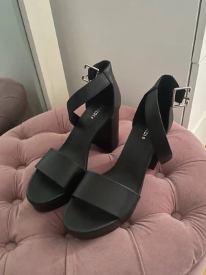Svarta pumps från Divided med klack - Stilrena svarta pumps från H&M med bred rem över tårna och justerbar rem med silverfärgat spänne runt ankeln. Skorna har hög, bred klack och är tillverkade i syntetmaterial. Perfekta för dig som vill ha en enkel men trendig look. Aldrig använda 