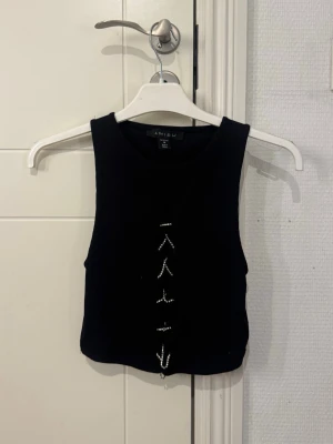 Svart croppad linnetopp  - Svart croppad linnetopp från Amisu med pärldetaljer framtill. Toppen är ärmlös och har en tight passform, perfekt att styla med höga jeans eller kjol. Materialet är ribbat och ger en snygg struktur. Storlek M, sitter som S