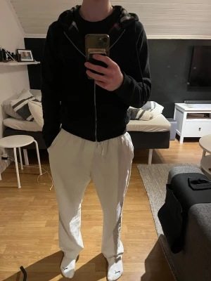 Svart hoodie från Burberry - Svart hoodie med dragkedja från Burberry. Klassisk rutig huva med snören och två fickor framtill. Perfekt för en chill och stilren look. Materialet är mjukt och bekvämt, och loggan är diskret broderad på bröstet. Tröjan har ett litet igensytt hål på högra armen. Ingenting man lägger märket till! Hör av dig vid fler frågor