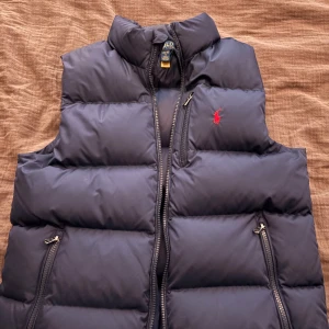 Mörkblå dunväst från Polo Ralph Lauren - Hej! Säljer en jättesnygg mörkblå dunväst från Polo Ralph Lauren med dragkedja och den ikoniska röda loggan på bröstet. Perfekt för tider som nu med kyliga dagar. Storlek 14-16 L men passar enligt mig för både S och M beronde på hur man vill ha den. Två praktiska fickor med dragkedja framtill. Riktigt bra skick!! Hör av dig om du har några frågor:)