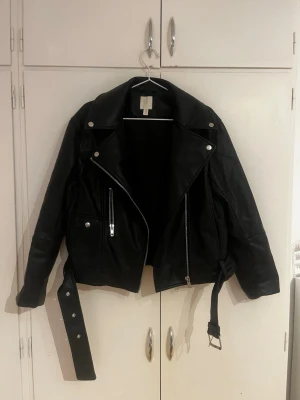 Svart biker skinnjacka med bälte - Snygg svart skinnjacka i klassisk bikerstil med bred krage, silverfärgade dragkedjor och knappar samt ett justerbart bälte i midjan. Jackan har lång ärm och coola detaljer som ger en edgy look. Perfekt för dig som vill ha en tidlös och trendig jacka.