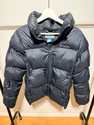 Svart pufferjacka från Columbia - Snygg svart pufferjacka från Columbia med hög krage och dragkedja framtill. Jackan har en quiltad design och är tillverkad i slitstark polyester som håller dig varm under kalla dagar. Perfekt för dig som vill ha en stilren och funktionell vinterjacka.