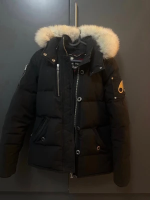Svart dunjacka med pälskant från Moose Knuckles - Snygg svart dunjacka från Moose Knuckles med fluffig vit pälskant på huvan och flera dragkedjor. Jackan har en quiltad look, praktiska fickor och ikonisk logga på ärmen. Perfekt för kalla vinterdagar och riktigt varm. Kvitto finns 