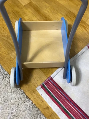 Blå gåvagn i trä - Säljer en klassisk gåvagn i trä med blå sidor och vita hjul. Vagnen har ett handtag i metall och en öppen låda för att lägga leksaker i. Perfekt för små barn som lär sig gå och vill köra runt sina grejer.