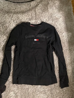 Svart sweatshirt Tommy Hilfiger - Snygg svart sweatshirt från Tommy Hilfiger med tryckt logga och flagga på bröstet. Rund halsringning och långa ärmar. Perfekt för en avslappnad och stilren look.
