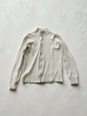 Beige stickad cardigan från These glory days - Beige stickad cardigan köpt fårn volt  med dragkedja framtill och långärmad design. Cardiganen  har en enkel, stilren look med ribbade muddar och krage. Perfekt för lager-på-lager och passar till många outfits.