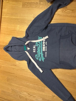 Blå Superdry hoodie med tryck - Snygg blå hoodie från Superdry med stort turkost och vitt tryck på bröstet samt japanska tecken. Tröjan har huva med vita dragsnören och en klassisk känguruficka framtill. Perfekt för en avslappnad och cool stil. Passar S-XS. Hör av er om pris och frågor. 