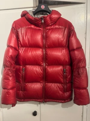 Röd pufferjacka med huva - Snygg röd pufferjacka från Colmar med glansig finish, huva och två dragkedjefickor framtill. Jackan har quiltad design och hel dragkedja. Perfekt för kalla dagar när du vill hålla stilen och värmen.
