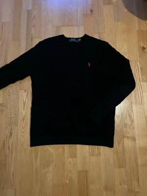 Svart kabelstickad tröja Polo Ralph Lauren - Klassisk svart kabelstickad tröja med mönster från Polo Ralph Lauren med röd broderad logga på bröstet. Rund halsringning och långa ärmar. Perfekt till lager på lager och funkar året runt. Snygg och tidlös design som alltid är rätt.