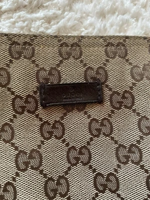 Gucci axelväska med GG-mönster - Snygg axelväska från Gucci med klassiskt GG-monogram i brunt på beige bakgrund. Väskan har en justerbar axelrem i grönt och rött tyg, samt en diskret läderdetalj framtill. Perfekt för dig som gillar ikoniska designeraccessoarer. Äkta kan visa kvitto till seriösa kunder.