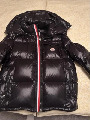 Svart dunjacka från Moncler - Snygg svart dunjacka från Moncler med glansig finish, huva och klassisk logotyp på bröstet. Jackan har en dragkedja med röd, vit och blå detalj längs öppningen. Perfekt för kalla dagar och riktigt stilren look.