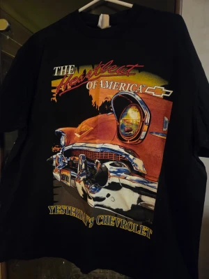 Svart vintage Chevrolet t-shirt - Cool svart t-shirt med färgglatt tryck av en klassisk Chevrolet och texten 'The Heartbeat of America' på framsidan. Perfekt för dig som gillar retrostil och bilar. T-shirten har normal passform och korta ärmar.