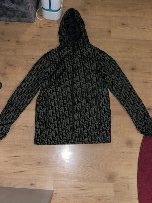 Svart vindjacka med mönster och huva - Snygg svart vindjacka med heltryckt mönster och huva. Jackan har dragkedja framtill och två sidofickor med dragkedja. Perfekt för dig som vill sticka ut med en unik look. Passar till både vardag och streetwear.