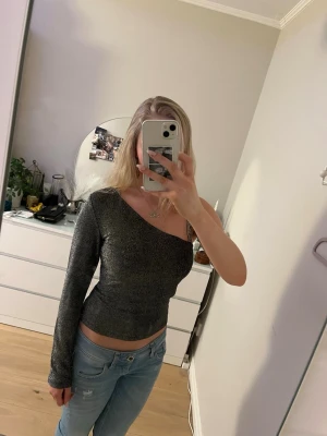 Glittrig svart one shoulder topp - Säljer en snygg svart glittrig topp med en axel och lång ärm. Tight passform och stretchigt material som ger en cool look. Perfekt för dig som vill sticka ut med en trendig och unik stil.