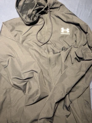 Svart vindjacka från Under Armour - Snygg svart vindjacka från Under Armour med huva och dragkedja framtill. Jackan har en sportig look, meshfoder i huvan och logga på bröstet. Perfekt för dig som gillar stilrena och funktionella plagg.