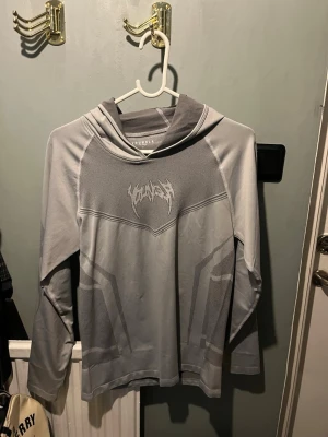 Grå compression hoodie från Youngla - Snygg grå compression hoodie från Youngla med tryck framtill i en futuristisk stil. Hoodien har långa ärmar, huva och är tillverkad i ett mjukt och stretchigt material. Perfekt för dig som gillar streetwear och stilrena plagg. Musclefit. Enbart provad 