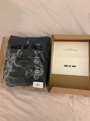 Svart hoodie från One of One - Svart hoodie med dragkedja från One of One. Framsidan har ett stort tryck med en ängel i strass och texten 'ONE OF ONE' i stora bokstäver. Hoodien har huva och ribbade muddar. Perfekt för dig som gillar unika streetwear-plagg. Den har en liten ”urtvättad” look men är fortfarande i fint och bra skick!