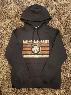 Mörkblå hoodie från Saint Laurent. Storlek S-M - Mörkblå hoodie från Saint Laurent med tryck i vitt och rött framtill. Klassisk huva med snörning och magficka. Perfekt för dig som gillar streetwear och vill ha en stilren look med exklusiv känsla.