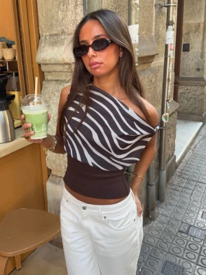Zebramönstrad oneshoulder topp - Snygg oneshoulder topp med zebramönster i svart och vitt. Toppen har en bred, ribbad midja i mörkbrunt och är croppad. Perfekt för dig som vill sticka ut med en trendig och unik look.