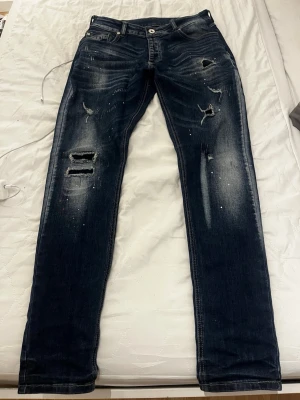 Mörkblå slitna jeans med raka ben - Säljer ett par mörkblå jeans med slitningar och hål på framsidan samt ljusa tvättade detaljer. Jeansen har klassisk femficksdesign och raka ben. Perfekta för dig som gillar en avslappnad och trendig look.