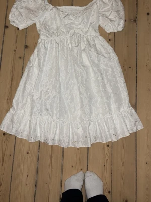 Vit klänning med puffärm och volang - Söt vit klänning med korta puffärmar och volangkant nertill. Klänningen har en romantisk känsla med broderade detaljer och smock upptill. Perfekt inför stundenten