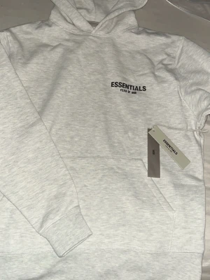 Ljusgrå Essentials Fear of God hoodie - Snygg ljusgrå hoodie från Essentials Fear of God med svart logga tryckt på bröstet. Klassisk huva och känguruficka framtill. Tillverkad i mjukt bomullsmaterial som känns skönt mot huden. Perfekt för en clean och avslappnad streetwear-look.