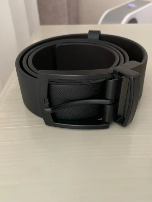 Svart stilrent bälte med metallspänne - Snyggt svart bälte i konstläder med matt svart metallspänne. Bältet har en enkel och modern design som passar till jeans eller kostymbyxor. Spännet har en diskret textur för extra stilpoäng.