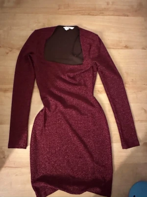 Vinröd glittrig kortklänning - Snygg vinröd kortklänning med glittereffekt och lång ärm. Klänningen har en unik fyrkantig urringning och figurnära passform. Perfekt för dig som vill sticka ut med en glittrig look. Materialet känns stretchigt och bekvämt. Pris kan diskuteras