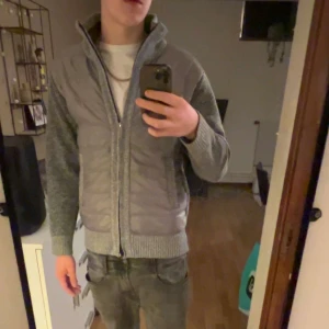 Cardigan - Säljer en grå cardigan jacka med stickade ärmar och quiltade partier framtill och baktill. Jackan har hög krage, ribbade muddar och dragkedja hela vägen. Perfekt för dagens väder!