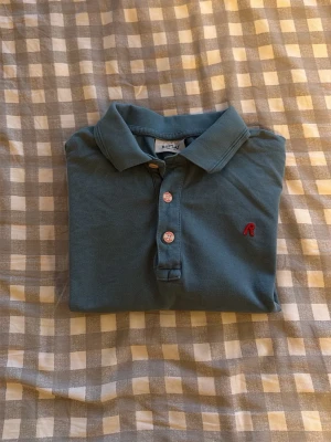 Blå pikétröja från Replay - Klassisk blå pikétröja från Replay med korta ärmar och krage. Tröjan har två vita knappar framtill och en liten röd logga broderad på bröstet. Priset går alltid att pruta