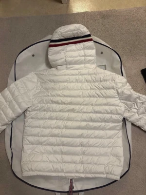 Vit dunjacka med huva och ränder - Snygg vit dunjacka med huva och horisontella quiltade sömmar. Huvan har en stilig röd, vit och marinblå rand. Jackan har långa ärmar med tryckknappar vid mudden och dragkedja framtill. Perfekt för kyliga dagar.