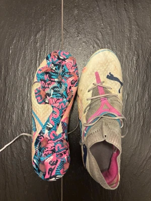 Puma future fotbollsskor - Säljer ett par vita Puma fotbollsskor med hög strumpa och snörning. Skorna har en färgglad sula i rosa, blått och svart mönster samt rosa och blå detaljer på ovandelen. Använda 3/4 gånger och lite smutsiga men hela och helt fungerande. Köpta för 2599.