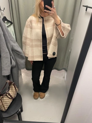 Beige rutig cape-kappa - Säljer en beige och ljusbrun rutig cape-kappa från Stylein. Kappan har vid passform och trekvartsärmar.. Tillverkad i ett mjukt ullblandat material och har en stilren look. Köpt på arkivet för 1500kr