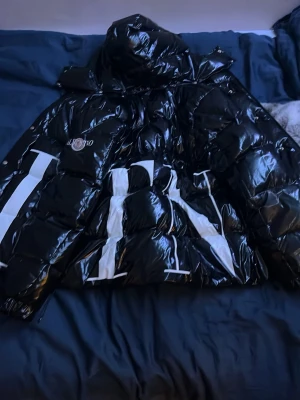 Svart pufferjacka från Moncler x Valentino - Snygg svart pufferjacka från Moncler x Valentino med stor vit logga framtill. Jackan har glansig finish, huva och dragkedjor på fickorna. Perfekt för dig som vill sticka ut med en exklusiv look och hålla dig varm under kalla dagar.