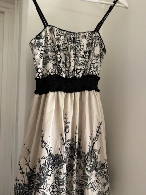 Vit och svart blommig klänning - Säljer en söt vit klänning från Vila med svarta blommiga mönster och smala axelband. Klänningen har en markerad midja med smock och är i kort modell. Perfekt för varma dagar och har en luftig känsla.