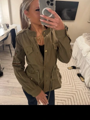 Olivgrön utilityjacka med snörning - Snygg olivgrön utilityjacka med snörning i midjan och dekorativa fickor framtill. Jackan har slitna detaljer på ena ärmen och stängs med knappar. Perfekt för dig som gillar en avslappnad och trendig stil.