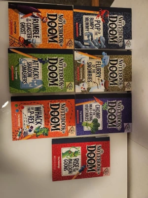 The Notebook of Doom bokserie - Sju böcker ur serien The Notebook of Doom av Troy Cummings. Omslagen är färgstarka med illustrationer av monster och äventyr. Böckerna är i pocketformat och har titlar som 'Rumble of the Coaster Ghost' och 'Rise of the Balloon Goons'. Perfekt för dig som gillar spännande och roliga berättelser.