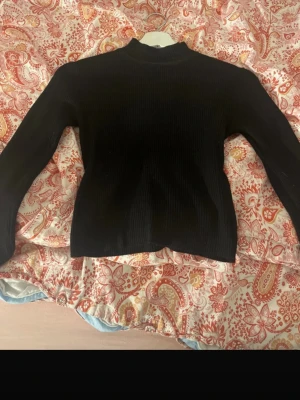 Svart ribbad stickad tröja - En svart, ribbad stickad tröja med lång ärm och hög hals. Tröjan har en enkel och stilren design som passar perfekt till jeans eller kjol. Materialet ser mjukt och stretchigt ut, vilket gör den bekväm att bära hela dagen.