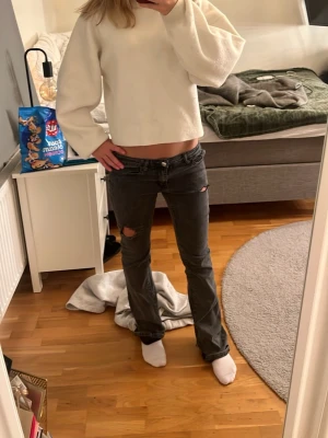 Grå slitna bootcut jeans - Säljer ett par grå jeans med bootcut-modell och slitningar vid knäna. Jeansen har låg midja och klassisk femficksdesign. Perfekta för dig som gillar en avslappnad och trendig stil.