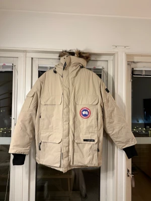 Beige parkas från Canada Goose - Säljer en beige parkas från Canada Goose med luva och äkta pälsdetalj. Jackan har flera praktiska fickor, svart mudd vid ärmslut och klassisk Canada Goose-logga på bröstet. Fodret är i dun. Perfekt för kalla dagar med sin rejäla och varma design. 