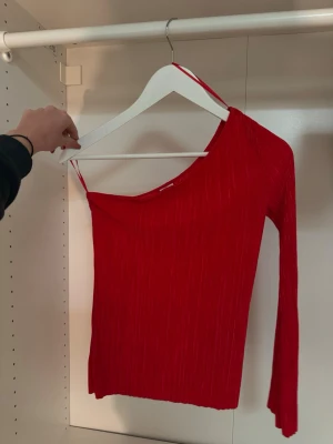 Röd oneshoulder topp från Gina Tricot - Trendig röd oneshoulder topp från Gina Tricot med ribbad struktur. Toppen har en lång vid ärm och en axelbandslös sida, vilket ger en cool asymmetrisk look. Perfekt för dig som vill sticka ut med en färgstark och modern stil.❤️