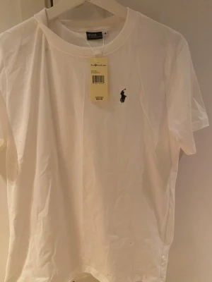Vit t-shirt från Polo Ralph Lauren - Klassisk vit t-shirt från Polo Ralph Lauren med rund hals och ikoniska broderade loggan på bröstet. Tillverkad i mjuk bomull och har en normal passform. Perfekt basplagg med stilren design. Priset är ganska fast priset kan diskuteras närmast hundringen 