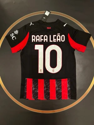 AC Milan matchtröja Rafa Leão - Snygg svart och röd AC Milan matchtröja med Rafa Leão och nummer 10 på ryggen. Tröjan har röda detaljer på ärmarna, vita Puma-loggor och klubbmärke. Perfekt för fotbollsfans som vill visa sitt stöd. S och L