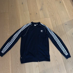 Adidas marinblå track jacket - Marinblå track jacket från Adidas med klassiska vita ränder längs ärmarna och vit logga på bröstet. Jackan har dragkedja framtill, ribbade muddar och är långärmad. Perfekt för en sportig och avslappnad stil.