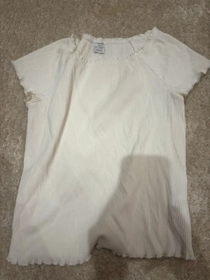 Vit ribbad topp från Lindex - Säljer en vit ribbad topp från Lindex med korta ärmar och lätt vågig kant nertill. Toppen har en enkel och clean look, perfekt att matcha med jeans eller kjol. Tillverkad i mjuk bomull för skön känsla hela dagen.