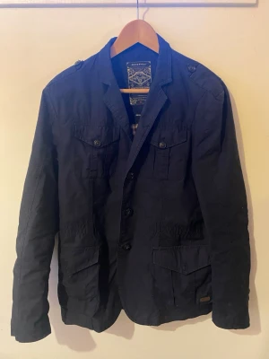 Field jacket från Reserved - Snygg mörkblå kavaj från reserved med fyra framfickor och knappar framtill. Klassisk krage, axelklaffar och detaljerade sömmar ger en cool och avslappnad look. Tillverkad i bomull för skön känsla och stilren passform.